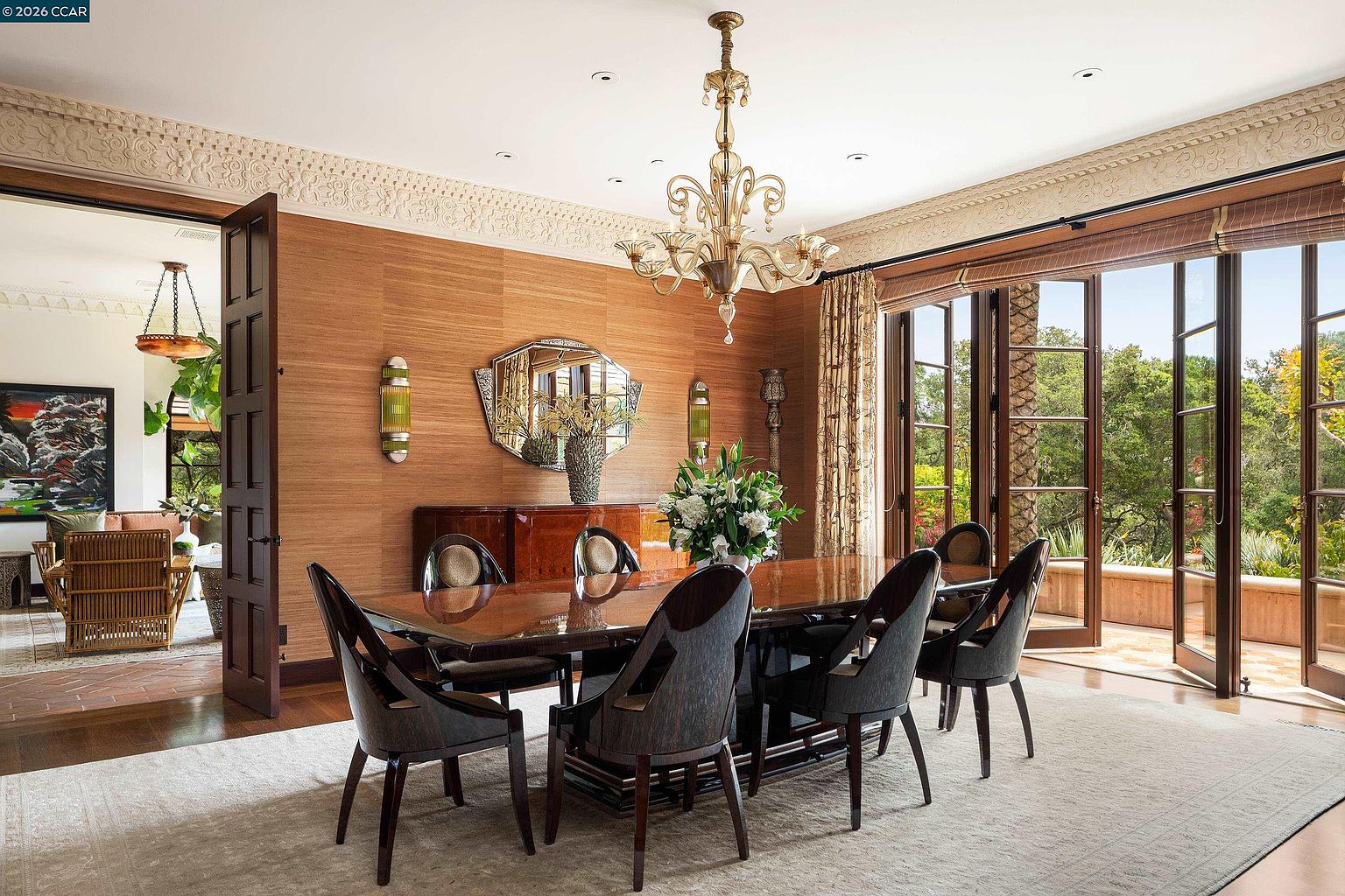 This elegant dining room features warm, textured wall coverings and a polished wood table surrounded by six dark, upholstered chairs. A grand chandelier serves as the focal point above the table, while large glass doors open to a lush outdoor view, flooding the space with natural light. The room is accented by a decorative mirror, wall sconces, and intricate crown molding, creating a sophisticated and inviting atmosphere.