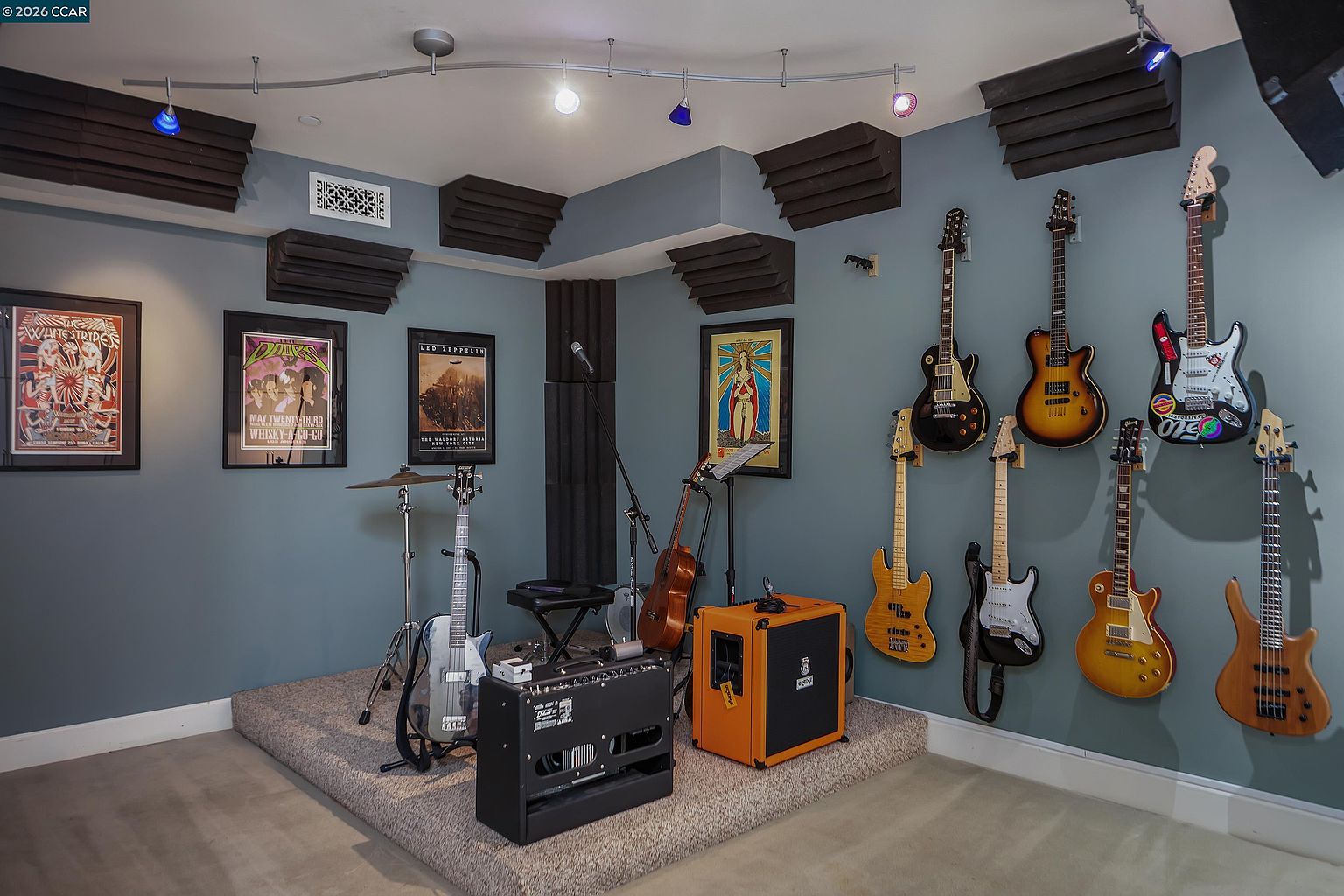 This dedicated music room features a raised carpeted platform designed for a drum kit and musical equipment, complete with acoustic foam panels mounted on the walls and ceiling corners. The space is decorated with framed rock music posters and a collection of electric and acoustic guitars mounted on the walls. The room is illuminated by a track lighting system, creating a focused and professional atmosphere for a home studio or practice space.