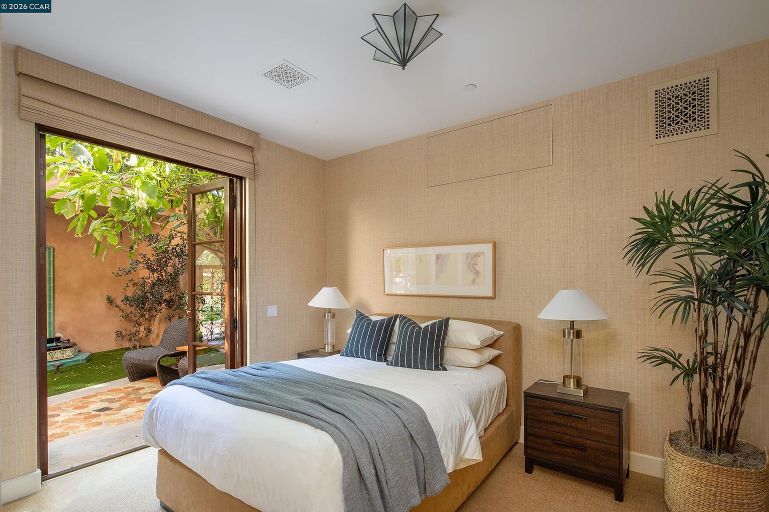 This serene bedroom features a neutral color palette with textured wall coverings and a comfortable bed dressed in white linens and a blue throw. Large glass doors open directly to a private, sunlit courtyard, seamlessly blending indoor and outdoor living spaces. A potted plant, matching bedside lamps, and a geometric ceiling fixture add a touch of modern elegance to the room.