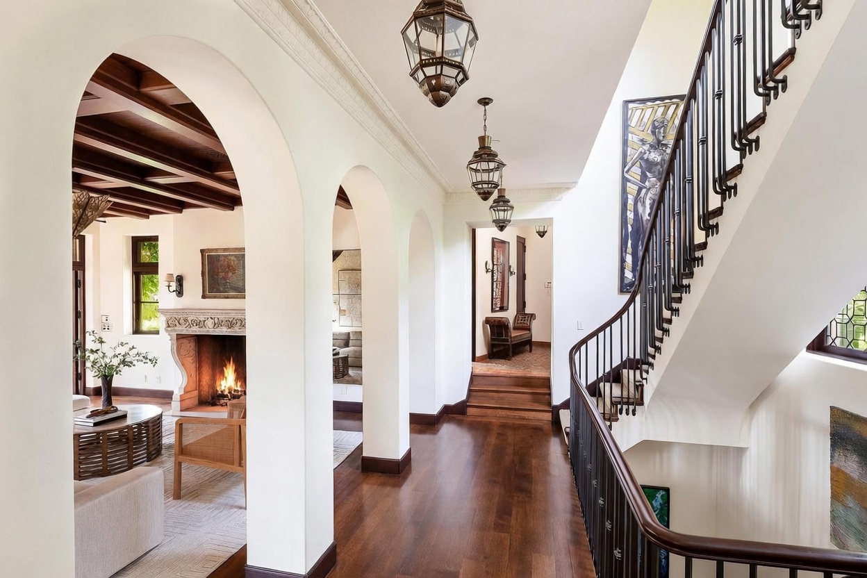 This elegant interior view captures a grand hallway featuring a series of white arched openings that lead into a living area with a stone fireplace and exposed wood ceiling beams. A sweeping staircase with dark metal railings ascends to the right, while three ornate, multi-tiered pendant lights hang from the ceiling to create a warm, sophisticated atmosphere. The perspective is from the top of the stairs, looking down the hallway, highlighting the architectural depth and the transition between the open living space and the private areas of the home.