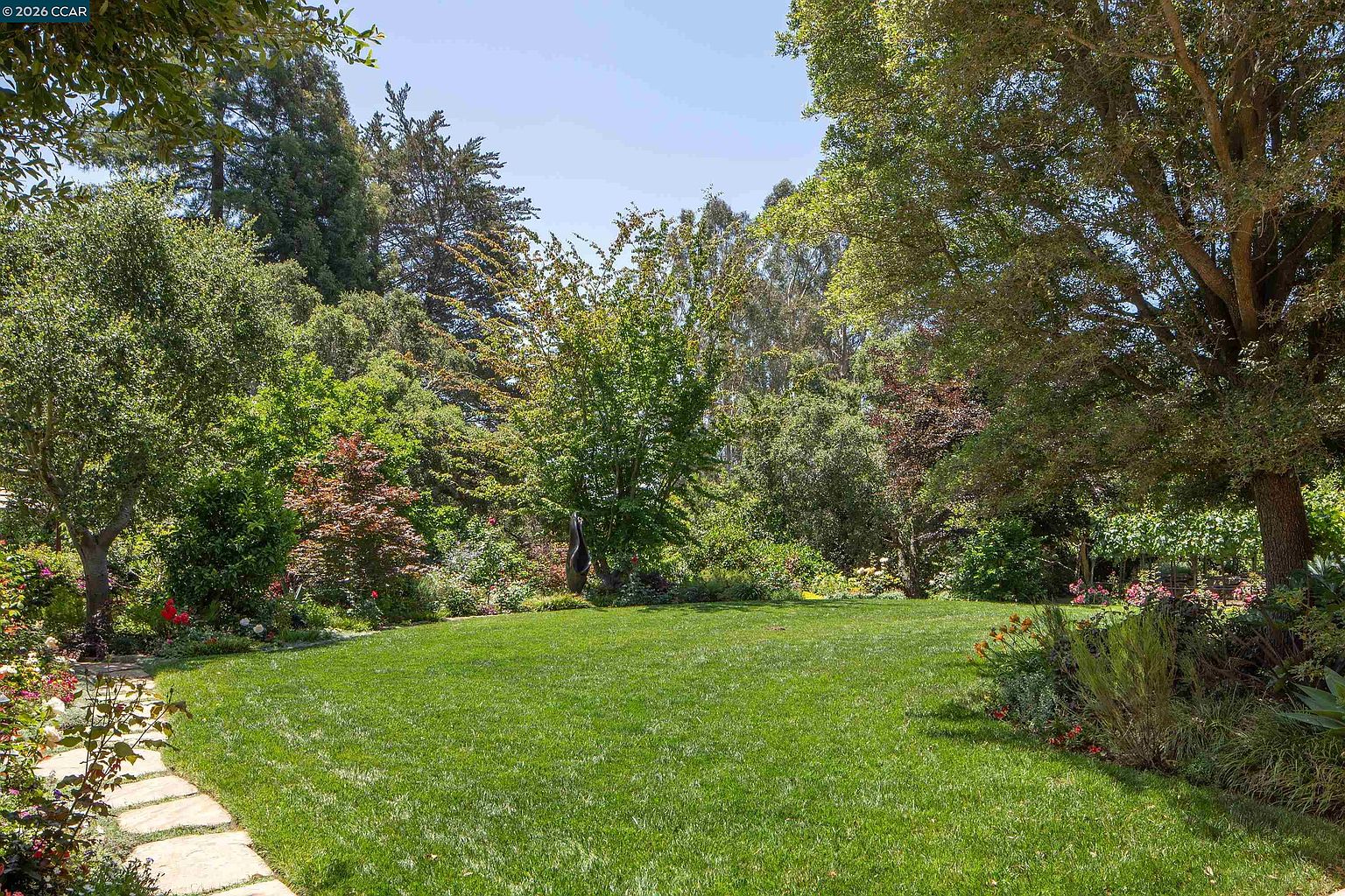 This lush, expansive backyard features a well-manicured green lawn framed by mature trees and vibrant garden beds. A stone pathway winds along the left side, leading toward a secluded area with a sculptural art piece nestled among the greenery. The scene offers a serene and private atmosphere, perfect for outdoor relaxation or garden enthusiasts.