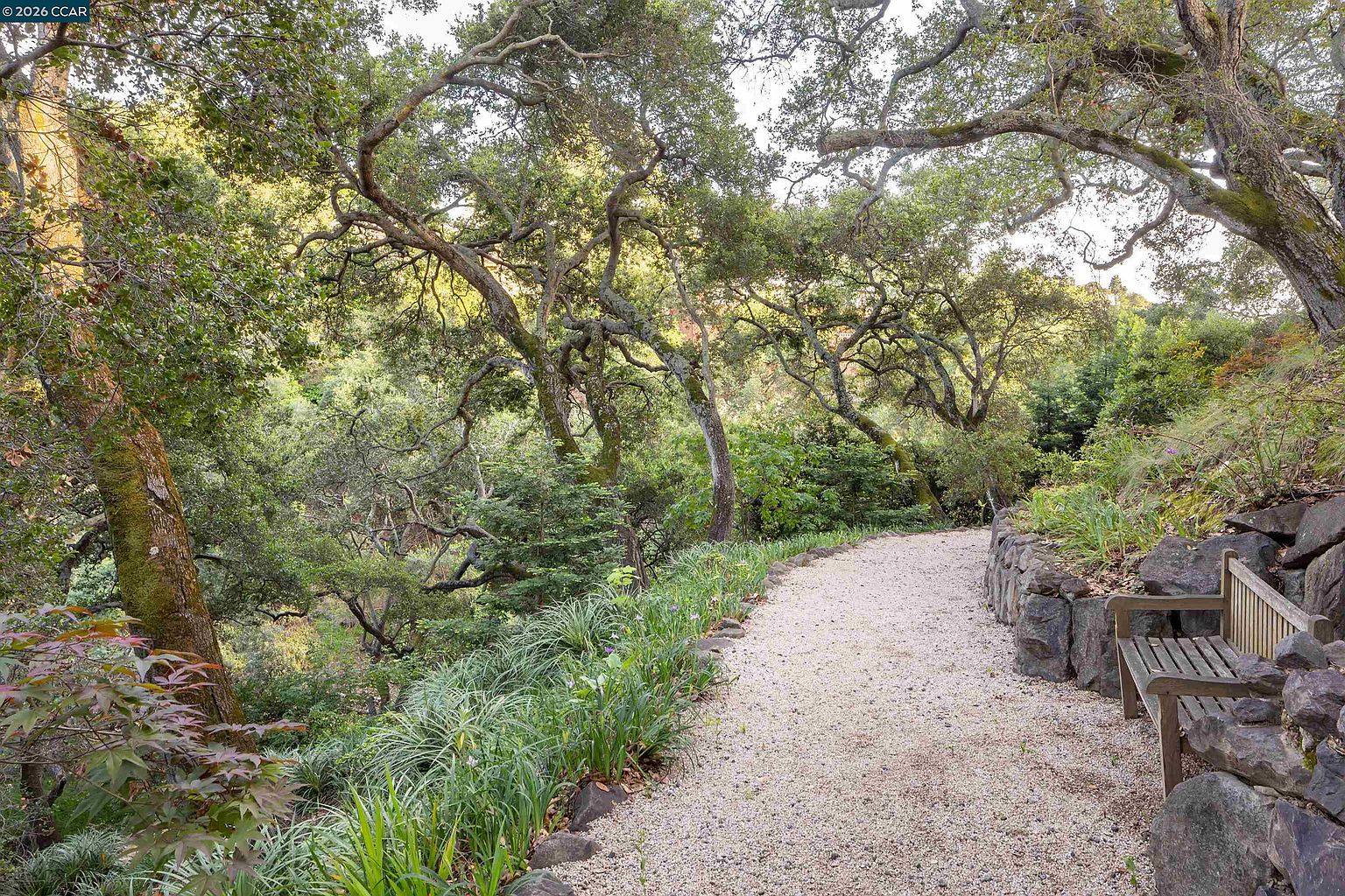 This serene garden path features a gravel walkway winding through a lush, wooded landscape filled with mature oak trees and dense greenery. A rustic wooden bench sits tucked against a stone retaining wall, offering a peaceful spot to enjoy the natural surroundings. The perspective captures the tranquil, park-like atmosphere of the property, emphasizing the integration of the home's outdoor space with the surrounding forest.