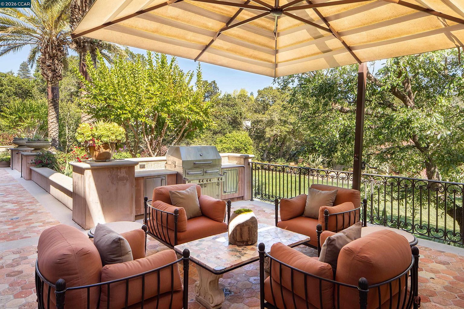 This inviting outdoor patio features a comfortable seating area with four plush orange armchairs arranged around a stone-topped coffee table. A built-in outdoor kitchen with a stainless steel grill and stone counter provides a functional space for entertaining, all shaded by a large, neutral-toned umbrella. The space is framed by a decorative wrought-iron railing that overlooks a lush, tree-filled backyard, creating a serene and private atmosphere.