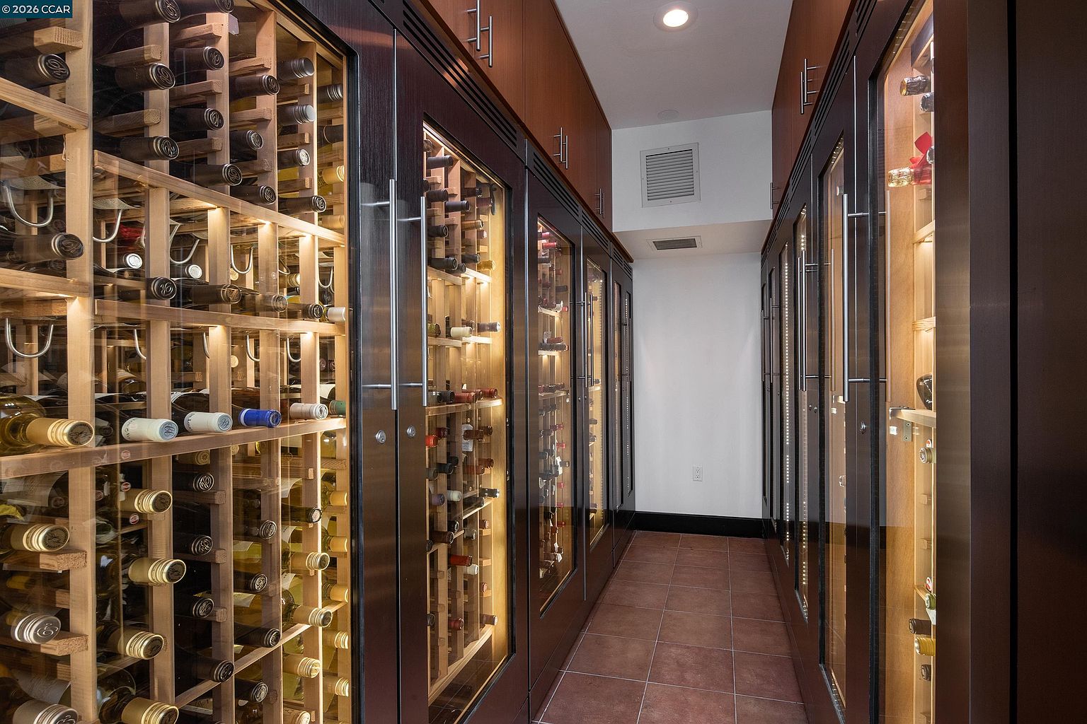 This sophisticated wine cellar features custom dark wood cabinetry with integrated glass-front doors and interior lighting, creating a gallery-like display for an extensive wine collection. The narrow corridor is lined with floor-to-ceiling wine racks on both sides, leading to a clean, white-walled end space with a ventilation vent. The floor is finished with warm-toned square tiles, and the overall aesthetic is one of luxury and organized storage.