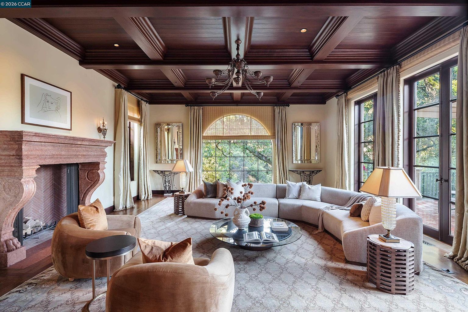This elegant living room features a coffered dark wood ceiling and a prominent stone fireplace with a decorative mantel. A large, curved sectional sofa and two plush armchairs are arranged around a circular glass coffee table, all set upon a patterned area rug. The room is bathed in natural light from large windows and glass doors that offer views of the surrounding greenery.