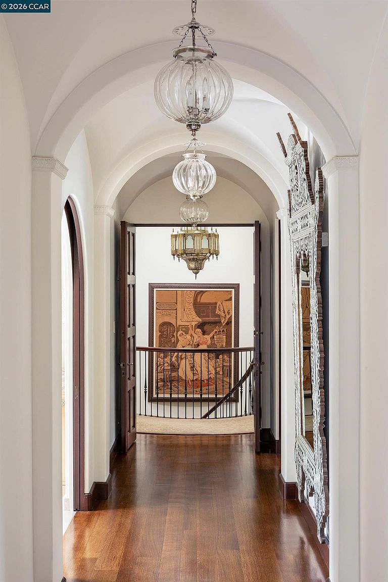 This elegant hallway features a barrel-vaulted ceiling and dark wood flooring, creating a sophisticated transition between spaces. A series of three glass pendant lights hangs in a row, leading the eye toward an open doorway that reveals a staircase with a decorative tapestry on the wall. To the right, a large, ornate mirror with a detailed frame adds a touch of grandeur to the corridor.