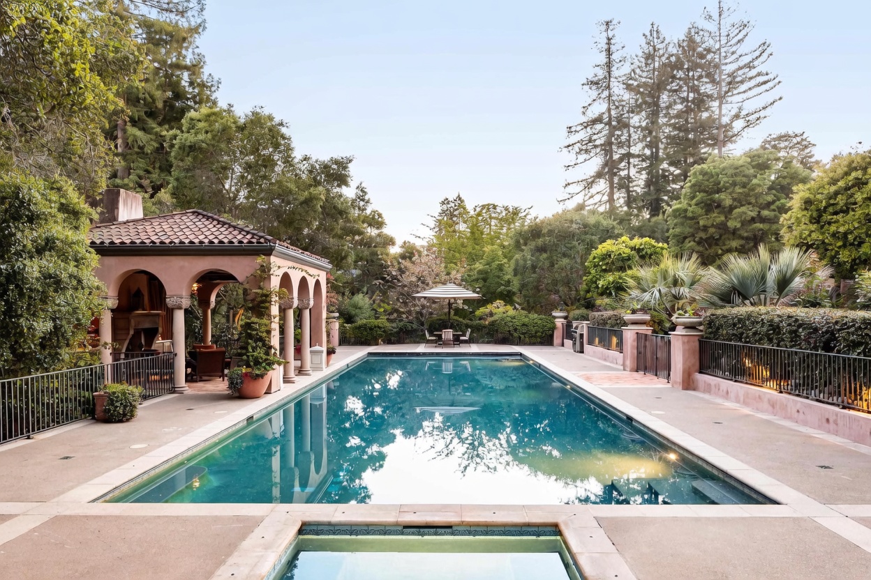 This elegant outdoor space features a long, rectangular swimming pool as the centerpiece, flanked by a paved deck and a covered loggia with arched openings. The area is surrounded by lush greenery and mature trees, creating a private, resort-like atmosphere. A small spa is visible in the foreground, while a dining set with an umbrella sits at the far end of the pool, enhancing the tranquil and sophisticated aesthetic.