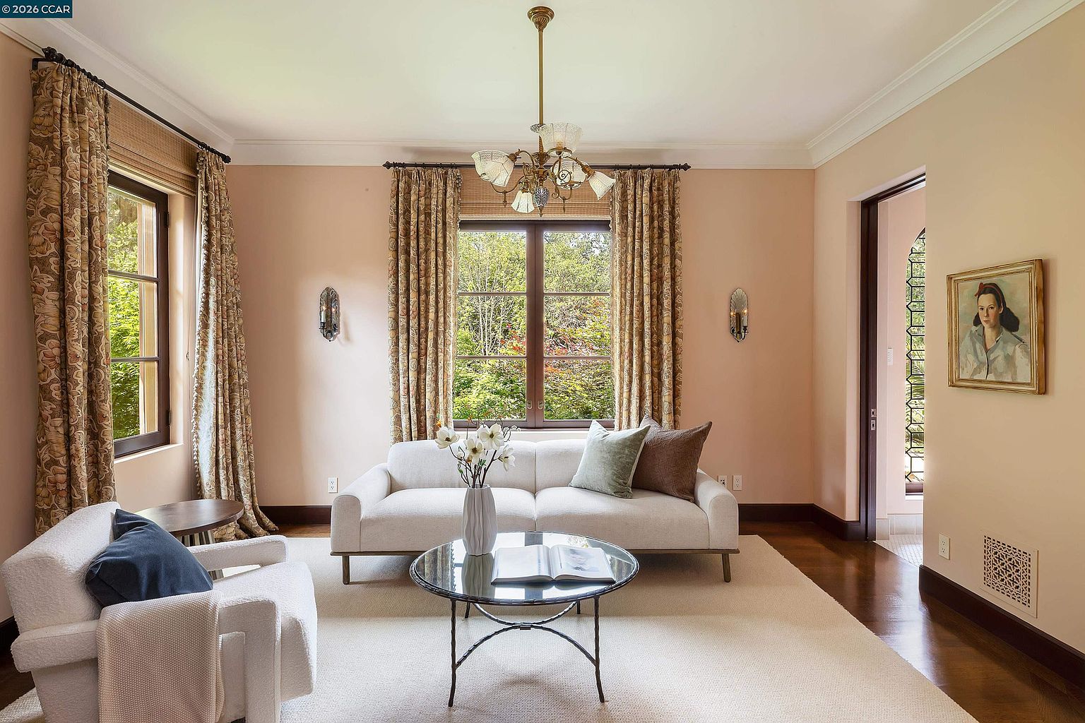 This elegant living room features soft peach-toned walls and warm hardwood flooring, creating a serene and inviting atmosphere. The space is anchored by a light-colored sofa and a matching armchair, complemented by a glass-topped coffee table and a central chandelier. Large windows draped in patterned curtains allow natural light to fill the room, offering a pleasant view of the lush greenery outside.