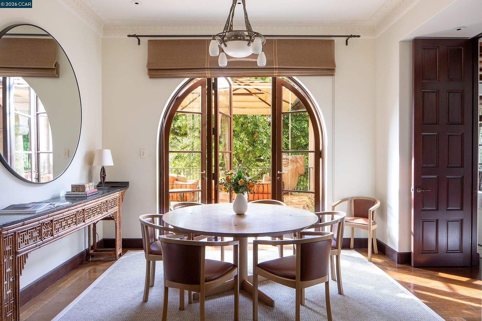 This elegant dining room features a central round wooden table surrounded by matching chairs, set atop a textured area rug. A large, circular mirror hangs above a decorative console table on the left, while a distinctive arched doorway with glass doors provides a view of the outdoor patio and lush greenery. The space is illuminated by a classic chandelier and benefits from natural light streaming through the arched entrance, creating a sophisticated and inviting atmosphere.