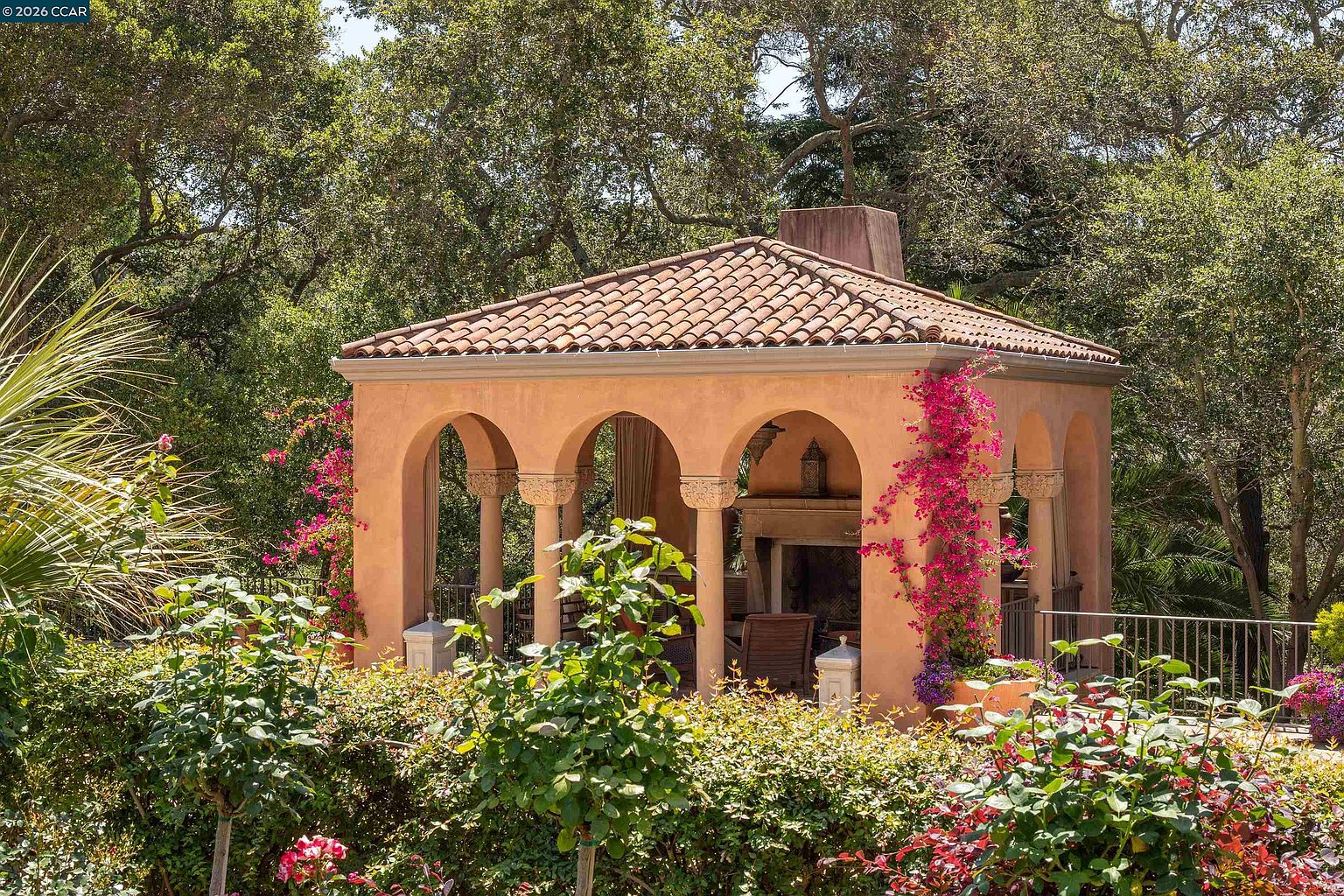 This image showcases a Mediterranean-style outdoor pavilion nestled within a lush, mature garden setting. The structure features arched openings, a terracotta-tiled roof, and a prominent stone chimney, surrounded by vibrant bougainvillea and dense greenery. The perspective captures the pavilion from a slightly elevated garden vantage point, emphasizing its role as a serene and elegant retreat.