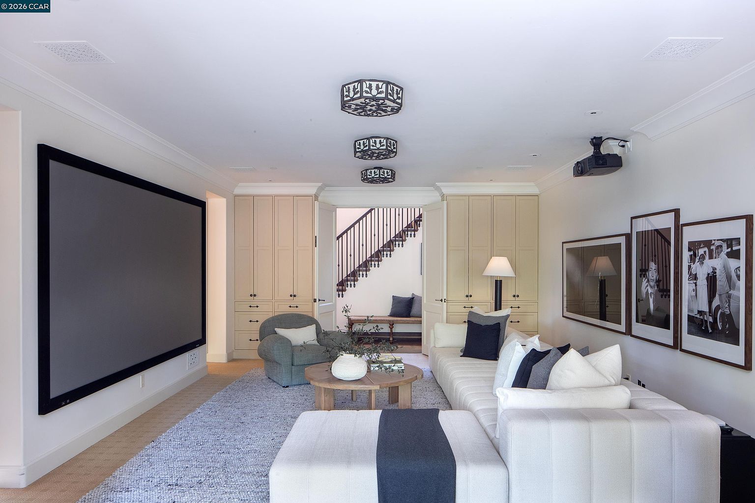 This sophisticated home theater room features a large projection screen, a plush sectional sofa, and a comfortable armchair, creating an inviting space for entertainment. The room is accented by three decorative circular ceiling fixtures and built-in cabinetry that frames a doorway leading to a staircase. The neutral color palette and soft textures contribute to a cozy, high-end atmosphere suitable for a media room.