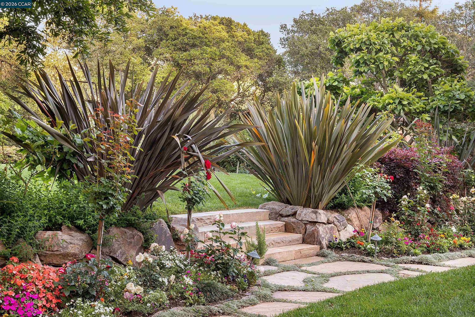 This lush, landscaped garden features a set of stone steps leading up to a grassy lawn, framed by two large, spiky ornamental grasses. The foreground is adorned with a vibrant mix of flowering plants and roses, while a stone path winds through the garden bed. The scene is set against a backdrop of mature trees, creating a serene and private outdoor retreat.