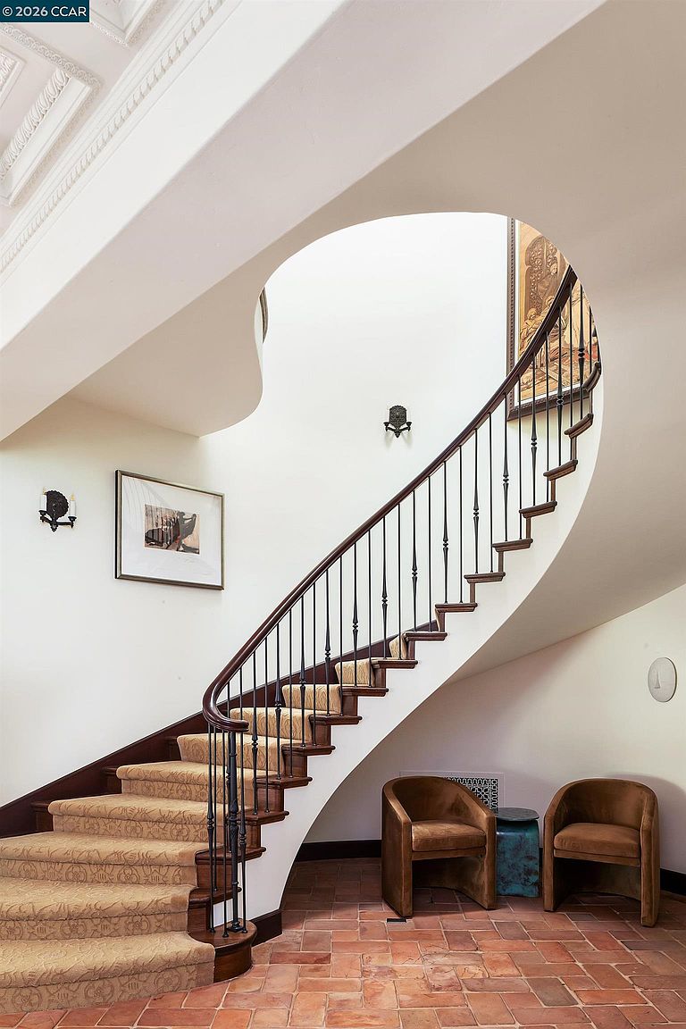This elegant entryway features a dramatic curved staircase with a dark wood handrail and ornate metal balusters, complemented by a patterned carpet runner. The space is anchored by terracotta-style floor tiles and two plush, curved armchairs positioned beneath the stairs. A framed piece of art hangs on the curved wall, enhancing the sophisticated, architectural character of the foyer.