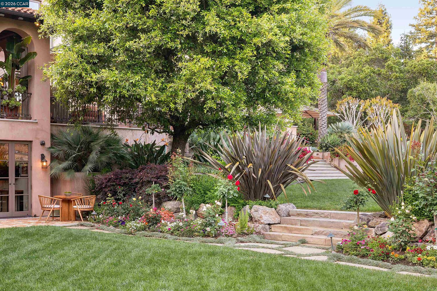 This lush, tiered backyard garden features a manicured lawn, stone retaining walls, and a variety of mature trees and ornamental plants. A small bistro set sits on a stone patio near the house, while stone steps lead up to a higher level of the landscape. The scene offers a serene, private atmosphere with a mix of vibrant flowers and structured greenery.