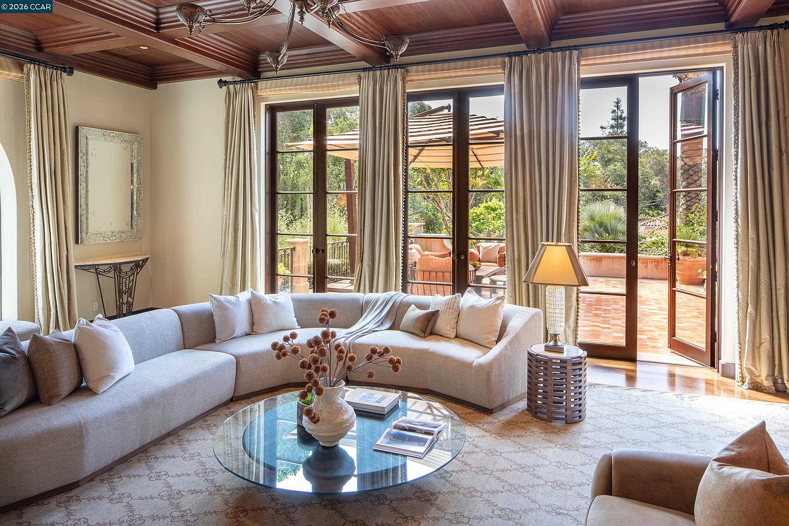 This elegant living room features a large, curved sectional sofa arranged around a circular glass-topped coffee table, creating a sophisticated seating area. The space is defined by a coffered wood-paneled ceiling and tall, multi-paned glass doors that open onto a sunlit patio, allowing for an abundance of natural light. A decorative mirror hangs above a console table, while a table lamp on a textured cylindrical base adds a warm, inviting glow to the room.