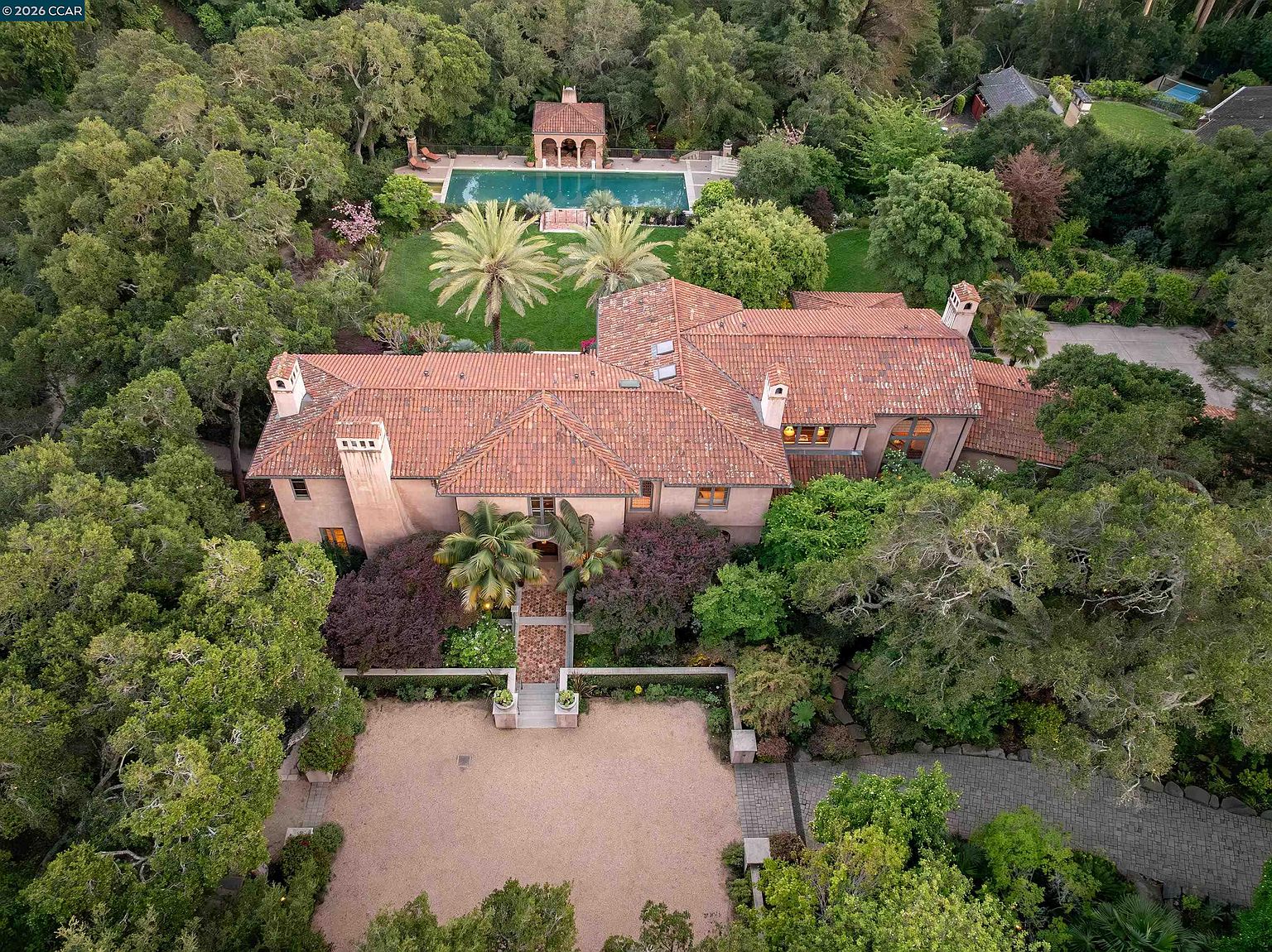 This high-angle aerial view captures a sprawling Mediterranean-style estate nestled within a lush, wooded landscape. The property features a prominent terracotta-tiled roof, a large gravel motor court at the front, and a beautifully landscaped backyard complete with a swimming pool and a classic pool house. The perspective highlights the expansive grounds, mature trees, and the elegant architectural symmetry of the residence.