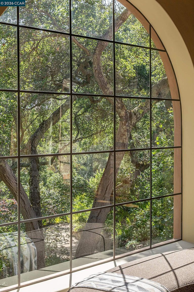 This image captures a large, arched window with a grid-patterned frame that serves as a focal point, framing a lush, wooded view outside. Below the window, a cushioned bench seat provides a cozy nook for relaxation, emphasizing the connection between the interior space and the natural surroundings. The perspective is intimate and focused, highlighting the architectural character and the serene, light-filled atmosphere of the room.