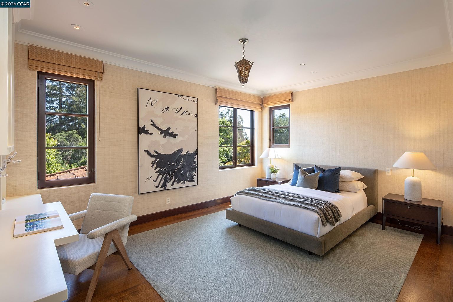 This bright and airy bedroom features neutral textured wallpaper, warm wood flooring, and a large area rug that anchors the space. The room is furnished with a queen-sized bed, two nightstands with matching lamps, and a dedicated workspace with a desk and chair. Large windows with woven shades allow for ample natural light, while a decorative pendant light and a large abstract art piece add character to the room.