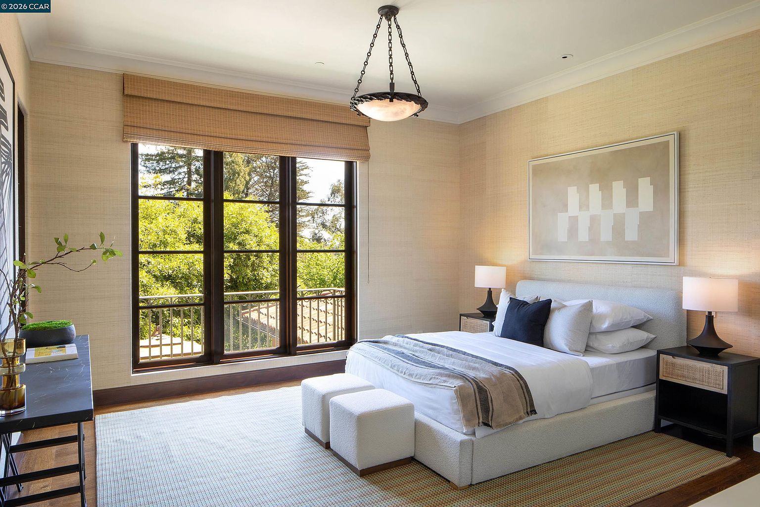 This bright and serene bedroom features textured wallpaper, a plush upholstered bed, and a pair of matching nightstands with modern lamps. A large window with woven shades provides ample natural light and a view of the lush greenery outside, while a unique pendant light fixture adds a touch of character to the space. The room is completed by a neutral-toned area rug and two small ottomans at the foot of the bed, creating a clean and inviting atmosphere.