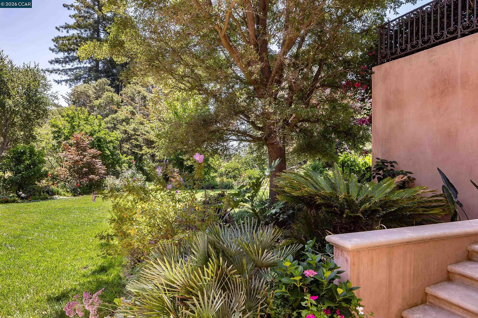 This lush, sun-drenched backyard features a well-manicured lawn bordered by mature trees and vibrant landscaping. A stone staircase leads up to a raised terrace area, which is enclosed by an ornate wrought-iron railing. The scene offers a serene and private atmosphere, perfect for outdoor relaxation or entertaining.