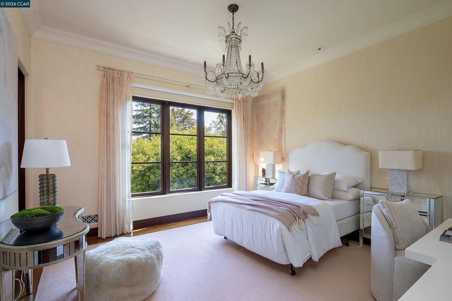 This elegant bedroom features a soft, neutral color palette with blush accents and a plush light-colored carpet. A central bed with a white upholstered headboard is flanked by mirrored nightstands and lamps, while a crystal chandelier adds a touch of sophistication above. Large windows with light curtains provide ample natural light and a view of the lush greenery outside, creating a serene and inviting atmosphere.