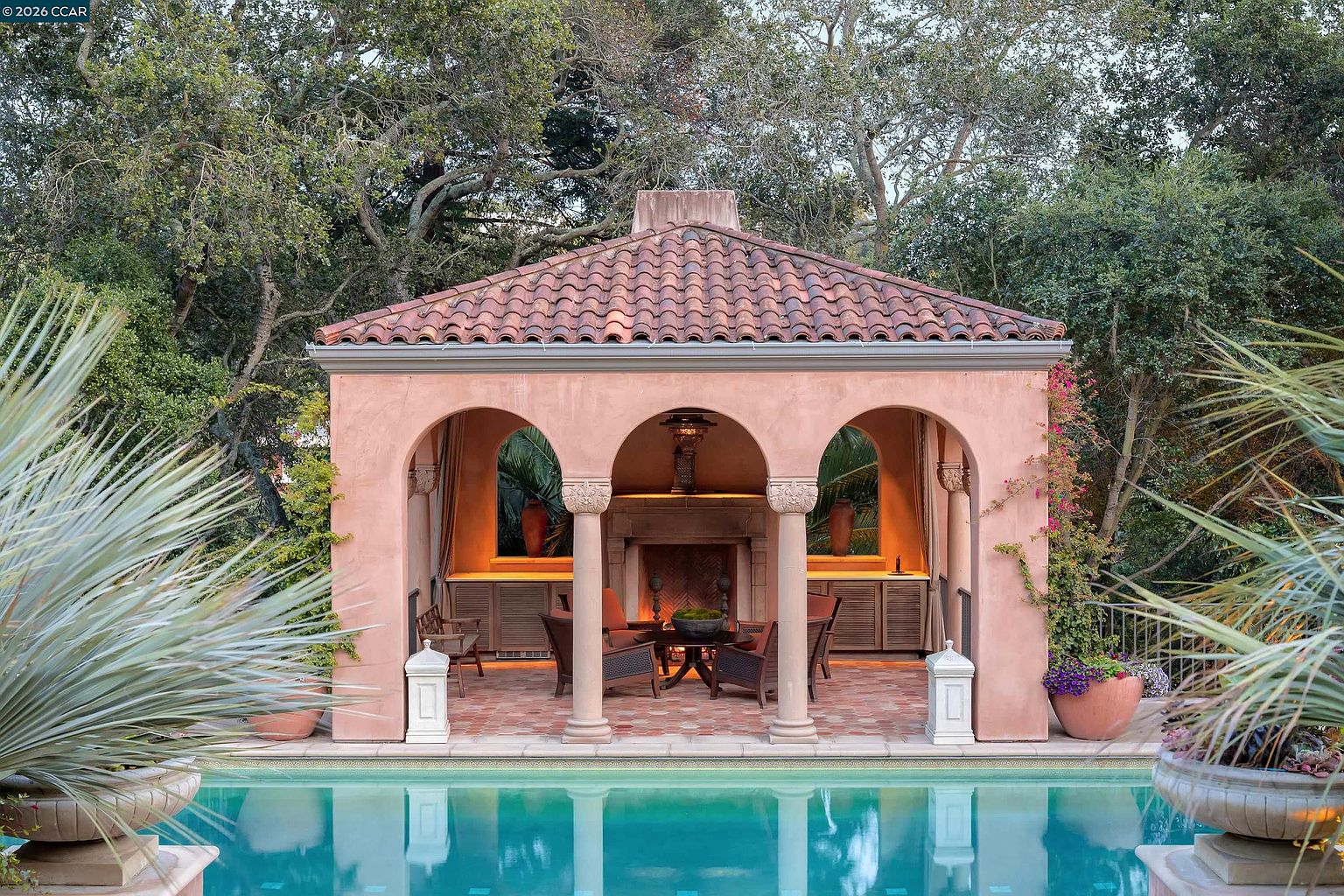 This elegant Mediterranean-style pool house features a terracotta-tiled roof and a symmetrical facade with three arched openings supported by stone columns. The open-air structure houses a cozy seating area centered around a stone fireplace, flanked by built-in cabinetry and warm accent lighting. The scene is framed by lush greenery and overlooks a serene swimming pool, creating a sophisticated and tranquil outdoor retreat.
