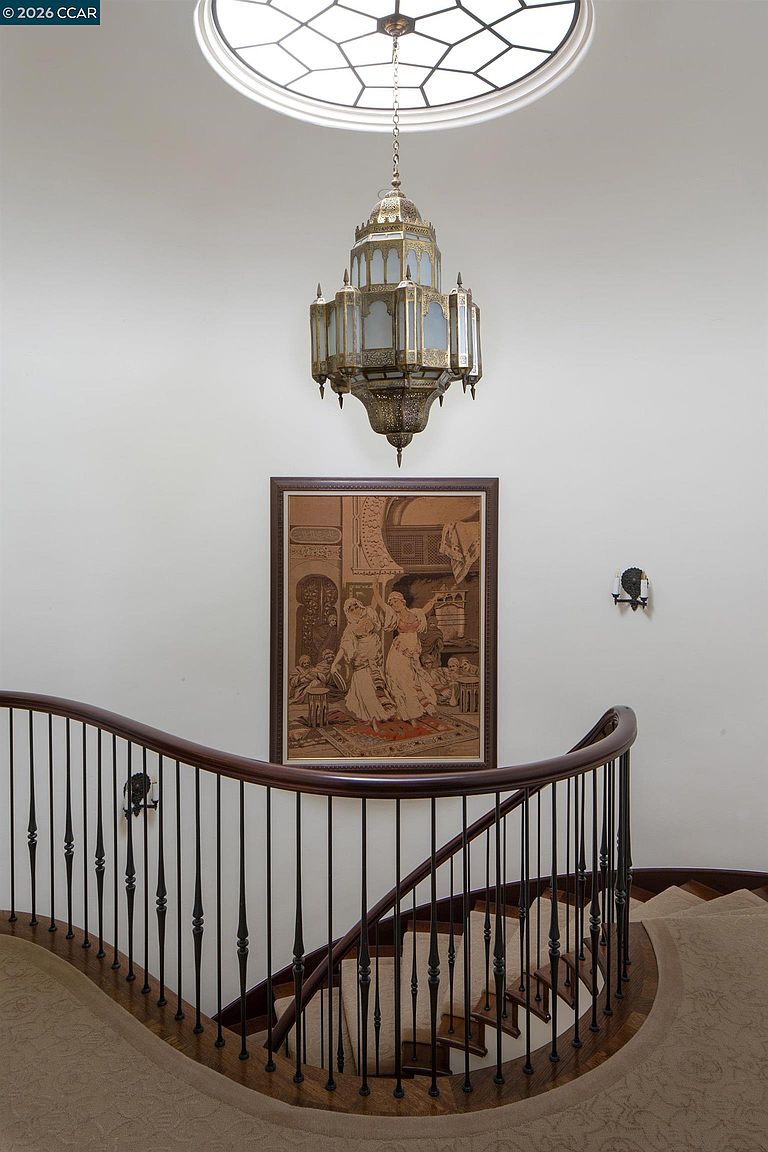 This elegant curved staircase features a dark wood handrail supported by slender black metal balusters, leading down to a carpeted landing. A large, ornate metal lantern chandelier hangs from a circular skylight above, casting light onto a framed piece of art depicting figures in a traditional setting. The perspective is centered and symmetrical, emphasizing the architectural grace of the curved staircase and the dramatic overhead lighting.