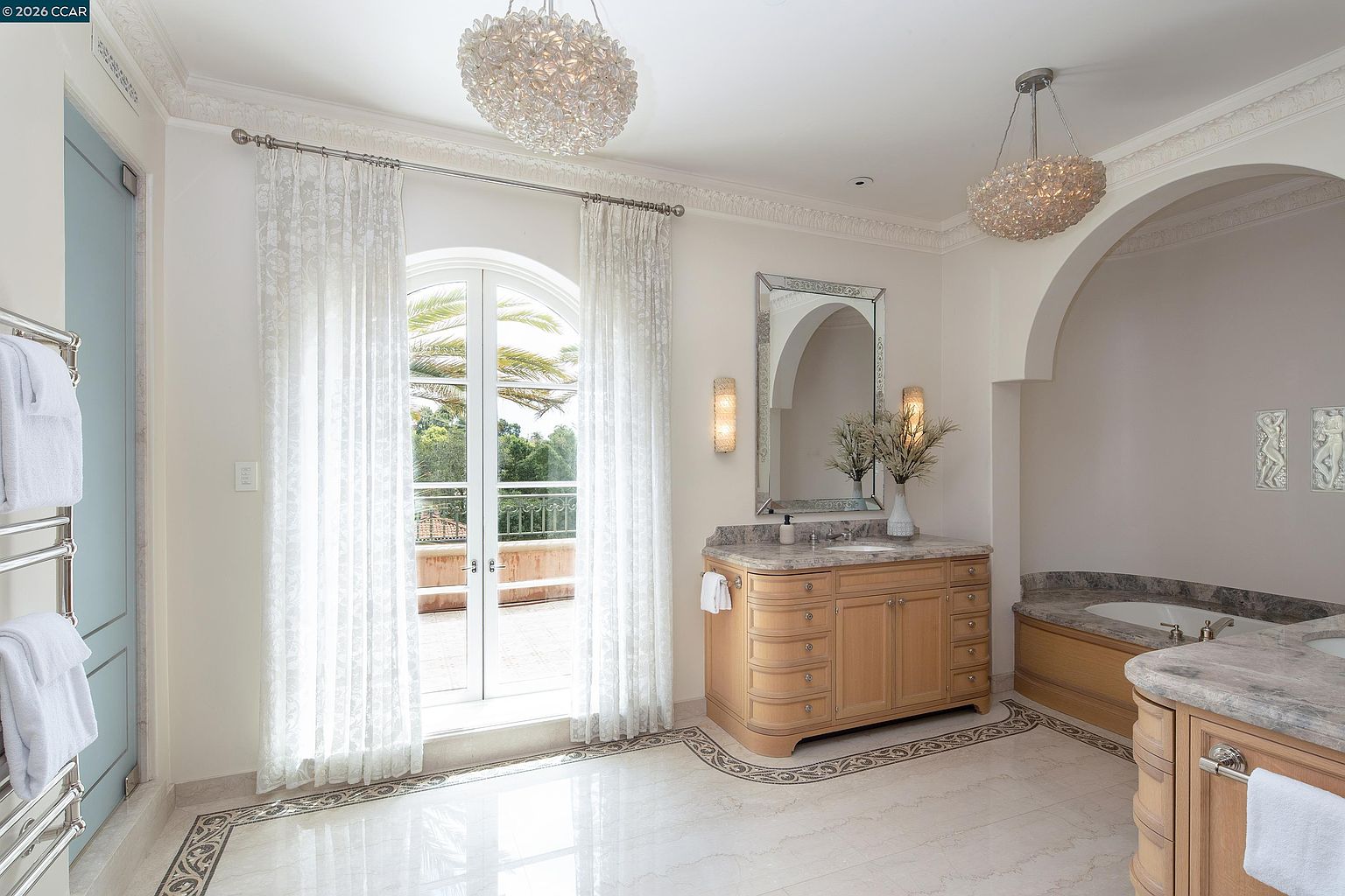 This elegant primary bathroom features a light wood vanity with a stone countertop, a large rectangular mirror, and two crystal-style chandeliers that add a touch of luxury. A set of French doors with sheer curtains opens onto a balcony, allowing natural light to fill the space, while a built-in soaking tub is tucked into an arched alcove. The room is finished with polished stone flooring featuring a decorative border, creating a sophisticated and serene atmosphere.
