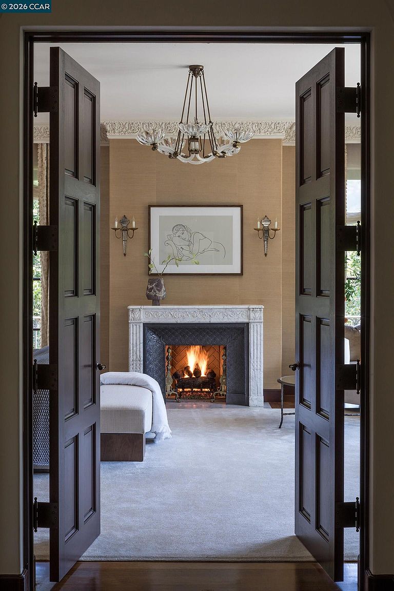 This elegant room is viewed through a set of dark, paneled double doors, creating a dramatic and inviting perspective. The space features a central fireplace with an ornate stone surround and a lit fire, flanked by wall sconces and topped with a framed piece of art. A chandelier hangs from the ceiling, and a neutral-toned chaise lounge sits in the foreground, contributing to a sophisticated and tranquil atmosphere.