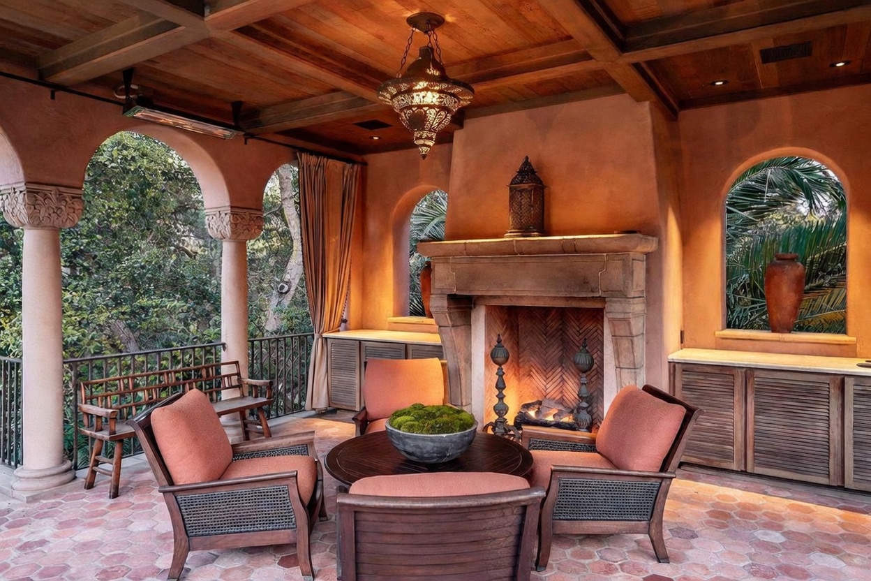 This elegant covered patio features a Mediterranean-inspired design with a stone fireplace as the central focal point. The space is furnished with comfortable seating arranged around a circular coffee table, set against a backdrop of arched openings and a wood-beamed ceiling. The warm, earthy tones of the terracotta-style flooring and stucco walls create an inviting, luxurious outdoor living area.