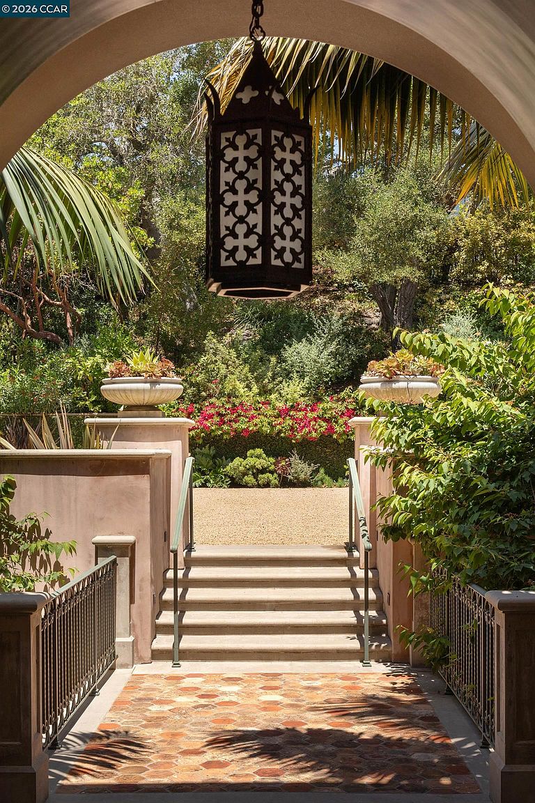 This elegant entryway features a terracotta-tiled landing framed by an arched stone opening, leading to a set of stone steps that descend into a lush garden. A decorative metal lantern hangs from the arch, while stone planters filled with succulents flank the staircase. The scene offers a Mediterranean-inspired aesthetic, emphasizing a seamless transition between the home's architecture and the vibrant, landscaped outdoor space.