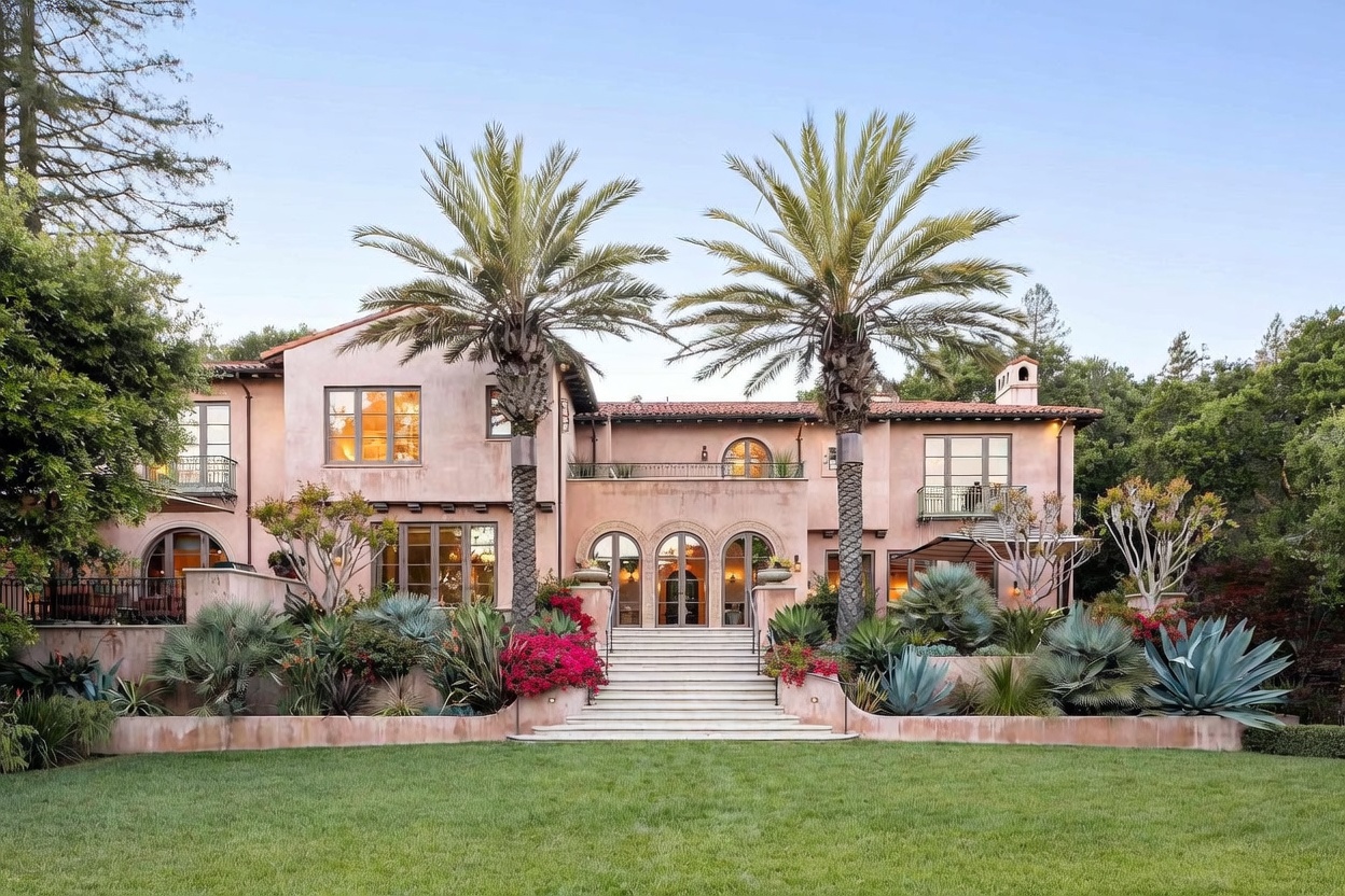 This Mediterranean-style estate features a warm, stucco-clad exterior with a terracotta tile roof and symmetrical architectural details. A grand set of stone steps leads from a manicured lawn up to a central arched entryway, flanked by two prominent palm trees. The rear facade includes multiple balconies with decorative iron railings and large windows that overlook the lush, tiered landscaping.