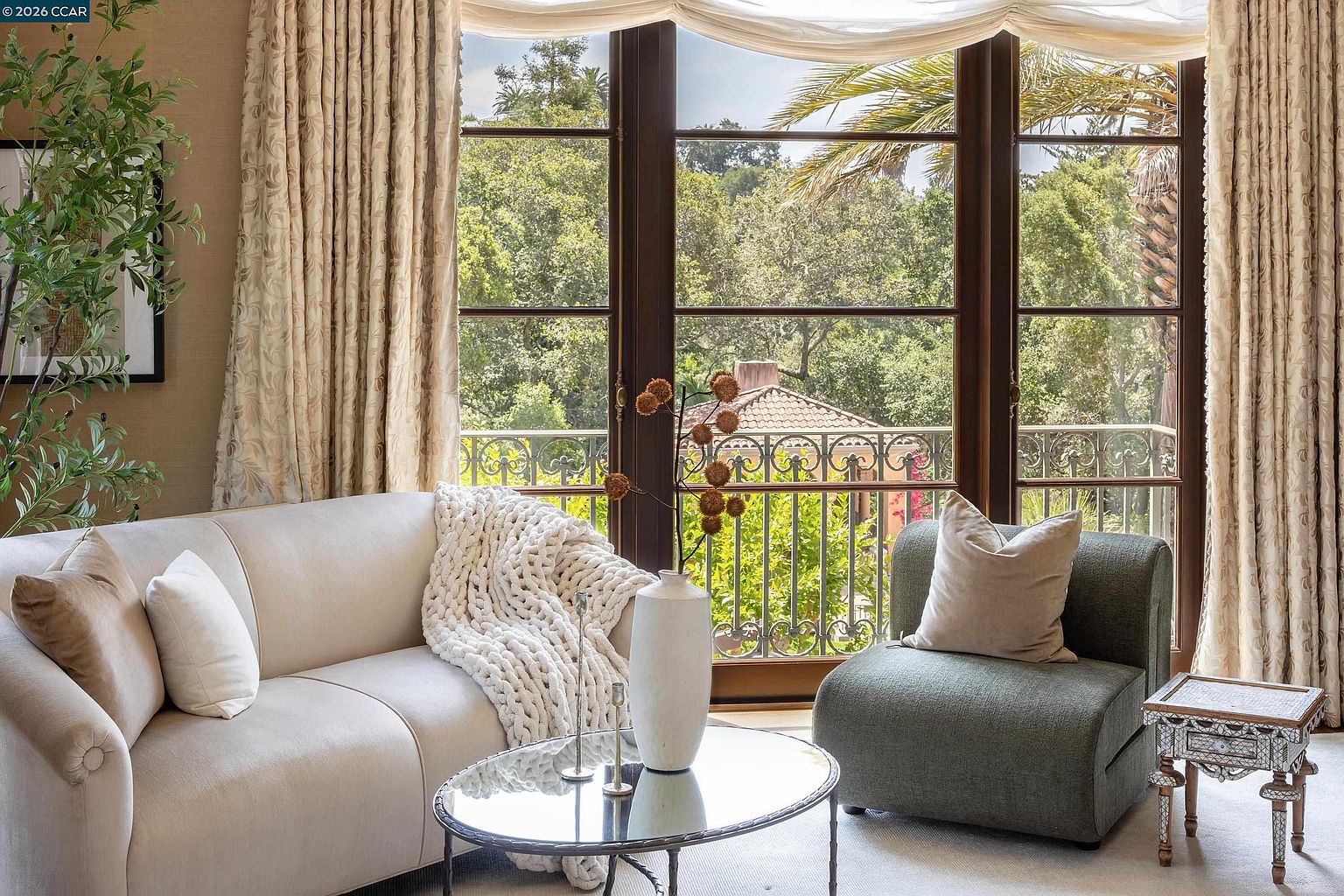This elegant living room features a neutral-toned sofa draped with a chunky knit throw, paired with a textured green accent chair and a glass-topped coffee table. Large windows with sheer drapery frame a scenic view of lush trees and a balcony with decorative iron railings. The space is accented by a tall vase with dried botanicals and a small, intricately detailed side table, creating a sophisticated and serene atmosphere.