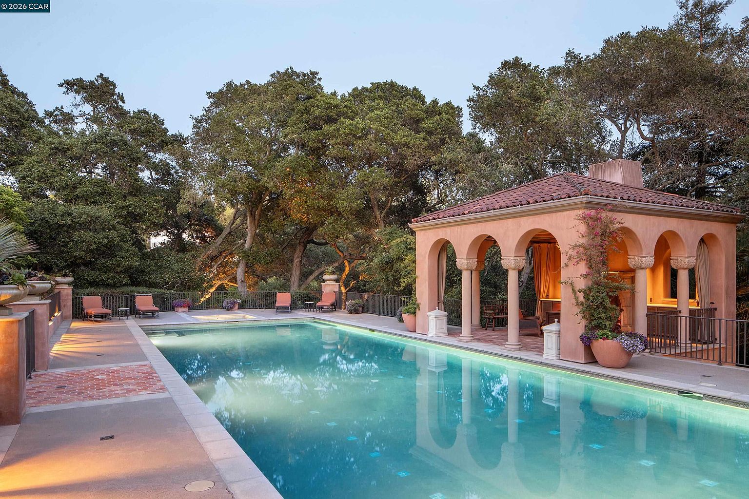 This serene outdoor space features a rectangular swimming pool set against a backdrop of lush, mature trees. To the right, a Mediterranean-style pool house with arched openings and a tiled roof provides a shaded lounge area, while a row of lounge chairs lines the pool deck. The scene is bathed in the warm, golden light of late afternoon, creating an inviting and tranquil atmosphere.