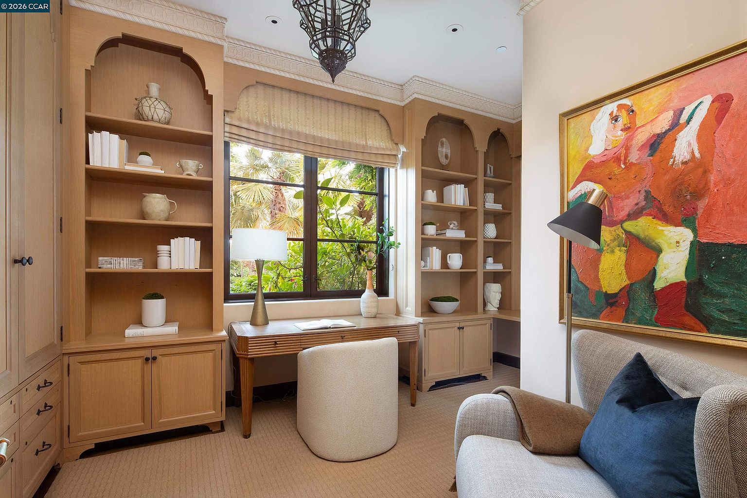 This elegant home office features custom light wood built-in shelving flanking a central window that overlooks lush greenery. A classic wooden desk is paired with a modern, curved upholstered chair, while a comfortable armchair with a navy blue pillow sits in the foreground next to a floor lamp. The room is accented by a large, vibrant abstract painting on the wall and a decorative chandelier, creating a sophisticated and tranquil workspace.