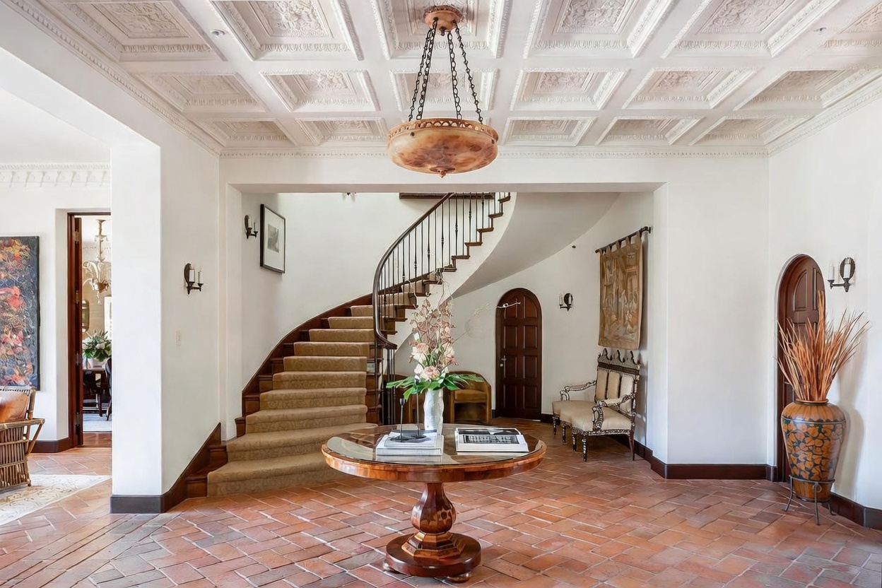 This grand foyer features a dramatic curved staircase with a wrought-iron railing and a central circular table topped with a floral arrangement. The space is defined by its intricate coffered ceiling, terracotta-style tile flooring, and arched wooden doorways that add a touch of Mediterranean elegance. The perspective is centered, capturing the architectural symmetry and the inviting, sophisticated atmosphere of the home's entrance.