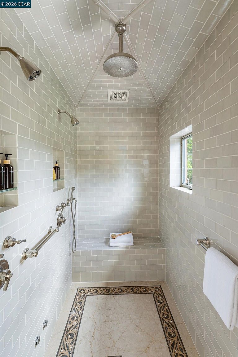 This spacious walk-in shower features light-colored subway tile on the walls and a vaulted ceiling, creating an airy and elegant atmosphere. The space includes a built-in bench, multiple shower heads, and a handheld sprayer, all finished in polished metal. A decorative patterned tile border frames the light-toned stone floor, while a small window provides natural light to the enclosure.