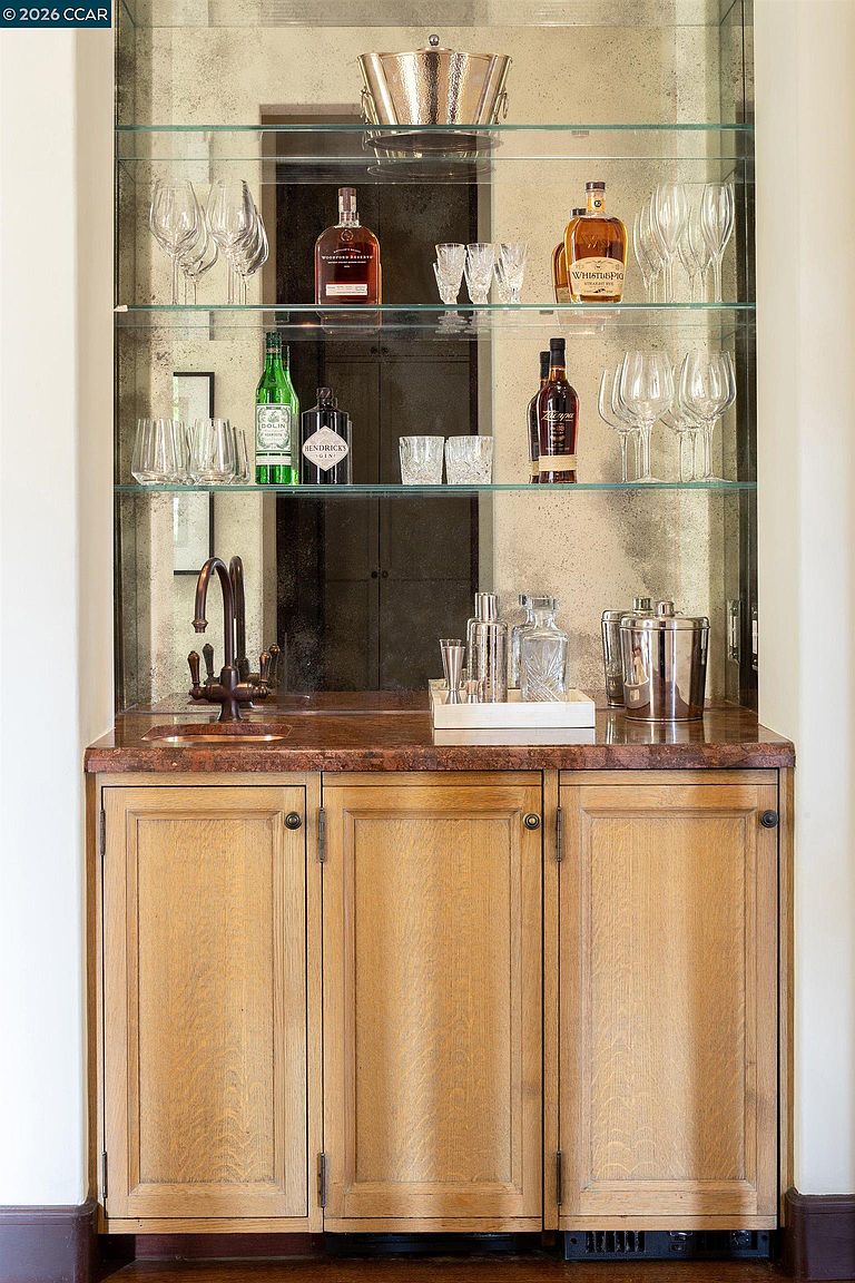 This image showcases a sophisticated built-in wet bar featuring light wood cabinetry and a rich, dark stone countertop. The space is accented by a mirrored backsplash and glass shelving that holds an assortment of glassware and premium spirits. A small, integrated sink with a dark metal faucet completes the functional design, creating an elegant area for entertaining.