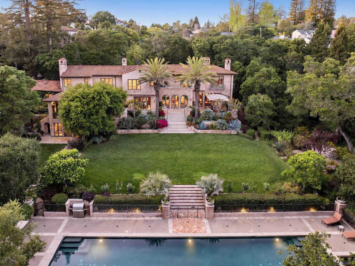 This elevated aerial perspective captures a grand Mediterranean-style estate featuring a sprawling manicured lawn, a large swimming pool in the foreground, and a multi-level residence with a terracotta tile roof. The property is surrounded by lush, mature trees, creating a private and serene atmosphere. Stone staircases connect the various levels of the landscape, emphasizing the elegant, tiered design of the outdoor living space.