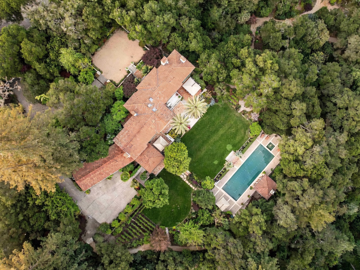 This high-angle aerial view captures a sprawling Mediterranean-style estate nestled within a lush, wooded landscape. The property features a large terracotta-tiled roof, a manicured central lawn, and a long rectangular swimming pool accompanied by a small pool house. A paved driveway leads to the residence, which is surrounded by dense mature trees, creating a private and secluded atmosphere.