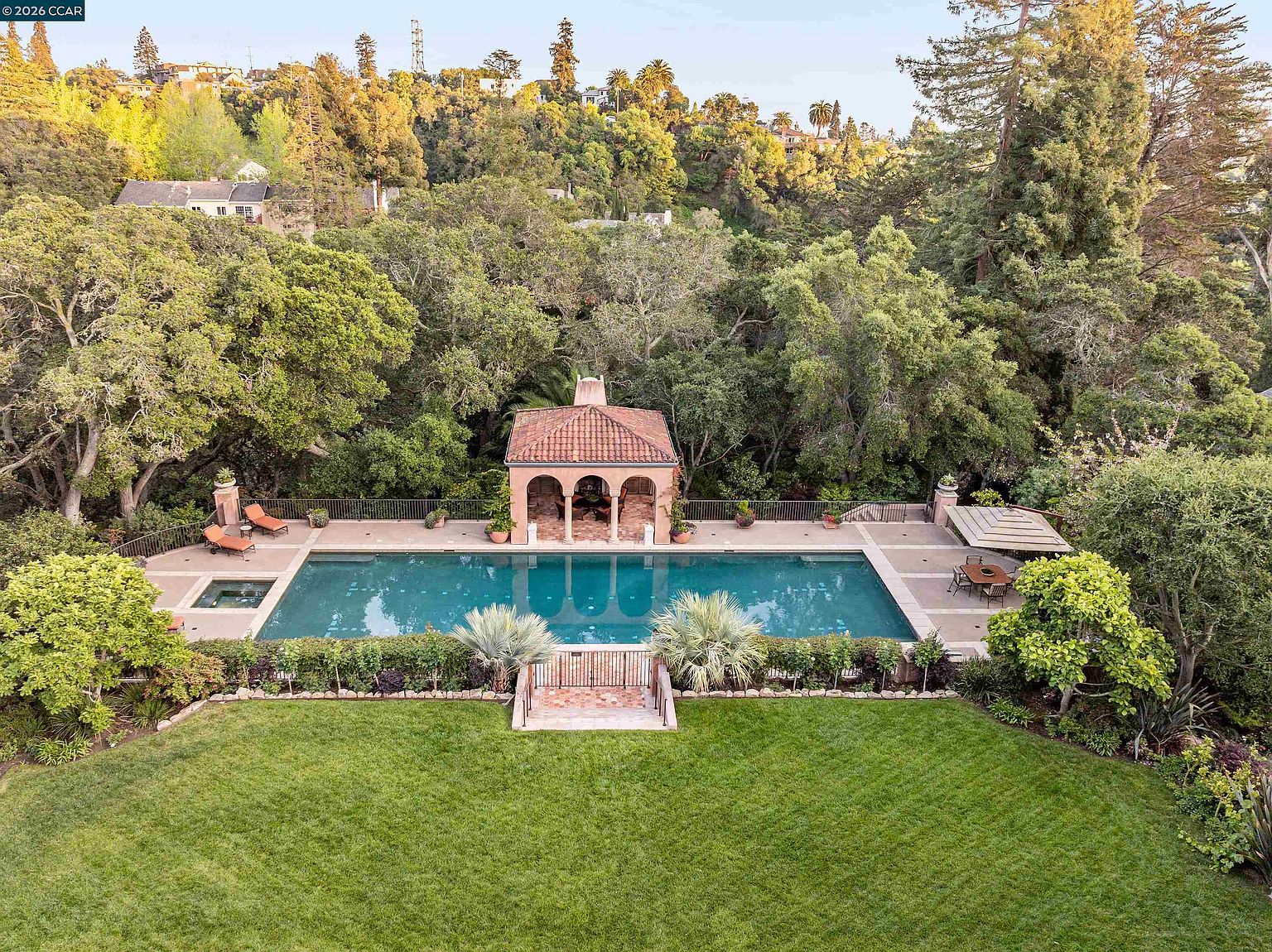 This elevated aerial view captures a luxurious backyard oasis featuring a large rectangular swimming pool and an integrated spa. A classic pool house with a tiled roof and arched openings sits centrally along the pool deck, flanked by lounge chairs and a shaded dining area. The property is surrounded by lush, mature trees and a manicured lawn, creating a private and serene outdoor retreat.
