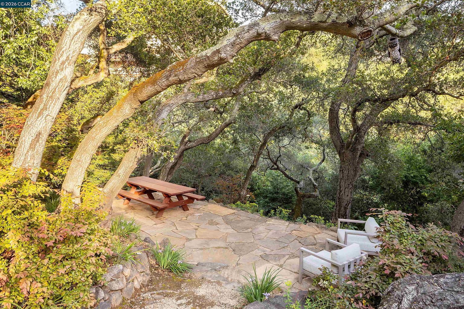 This serene outdoor patio features a natural stone flagstone surface nestled beneath a canopy of mature, sprawling oak trees. The space is furnished with a wooden picnic table and two comfortable lounge chairs, creating an inviting area for relaxation or al fresco dining. The surrounding lush greenery and dappled sunlight provide a peaceful, secluded atmosphere perfect for enjoying the natural landscape.