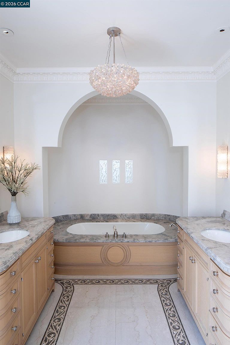 This elegant primary bathroom features a symmetrical layout with dual vanities flanking a central soaking tub set within a decorative arched alcove. The space is highlighted by a crystal chandelier, intricate patterned tile flooring, and light wood cabinetry topped with stone counters. The perspective is centered, emphasizing the sophisticated architectural details and the luxurious, spa-like atmosphere of the room.