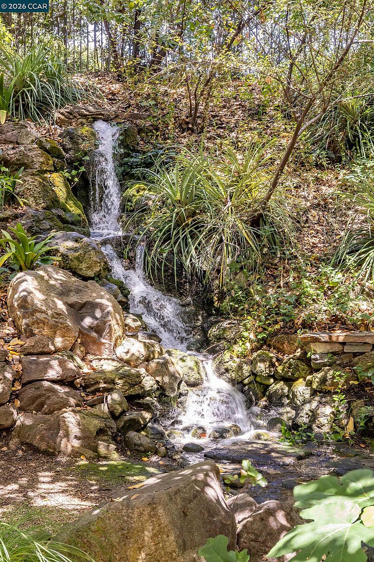 This serene garden feature showcases a tiered, naturalistic stone waterfall cascading into a small, rocky stream. The landscape is lushly planted with various grasses and shrubs, creating a tranquil, woodland-inspired atmosphere. The perspective is a close-up, eye-level shot that emphasizes the texture of the mossy rocks and the movement of the water.