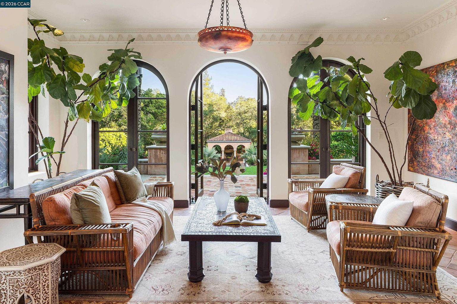 This elegant living room features a symmetrical layout with rattan-style seating arranged around a central patterned coffee table. Three large arched glass doors provide a seamless transition to the outdoor garden, allowing natural light to flood the space. The room is accented by tall indoor trees and a decorative hanging pendant light, creating a sophisticated and airy Mediterranean-inspired atmosphere.