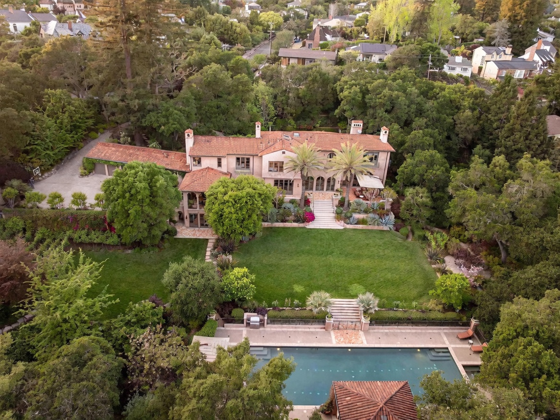 This high-angle aerial view captures a sprawling Mediterranean-style estate nestled within a lush, tree-filled landscape. The property features a large, multi-level residence with a terracotta tile roof, a manicured lawn, and a prominent swimming pool with an adjacent pool house. The perspective highlights the expansive grounds, mature landscaping, and the seamless integration of the home into its private, wooded surroundings.