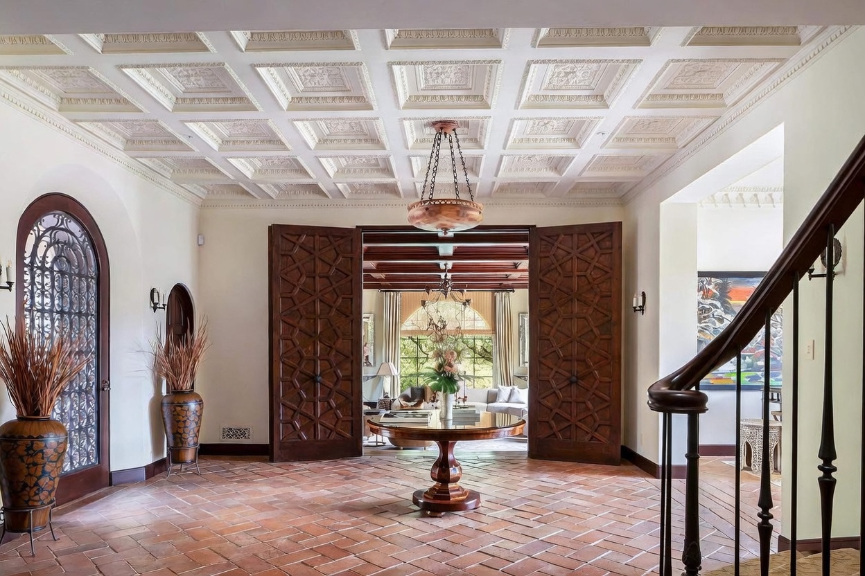 This grand foyer features a striking coffered ceiling with intricate decorative molding and a central pendant light. The space is anchored by a circular wooden table on a terracotta-tiled floor, framed by ornate, dark wood double doors that lead into an adjacent living area. To the left, a tall arched glass door with iron detailing provides a view of the exterior, while a wooden staircase with a decorative railing ascends on the right.