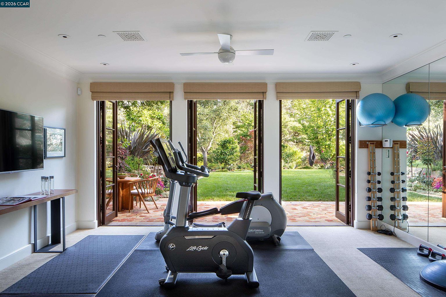 This bright and airy home gym features a stationary exercise bike as the central focus, set upon a dark rubberized mat. The room is framed by three sets of French doors that open onto a lush, green backyard, providing an abundance of natural light and a serene view. To the side, a mirrored wall reflects the space, which also includes a wall-mounted rack of dumbbells and two large exercise balls, creating a functional and inviting workout environment.