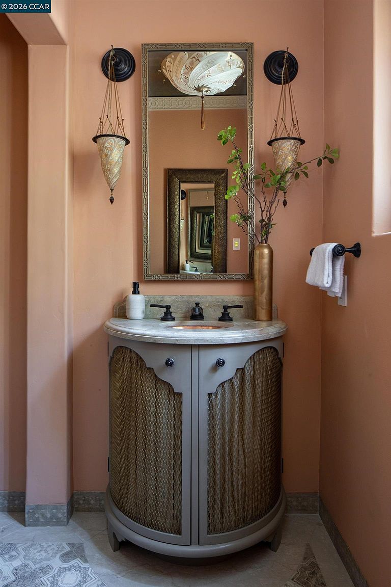 This elegant powder room features a unique curved vanity with a textured mesh front and a stone countertop. The space is accented by warm, peach-toned walls, a decorative framed mirror, and two ornate hanging light fixtures. A tall gold vase with greenery adds a touch of organic texture to the sophisticated, eclectic design.