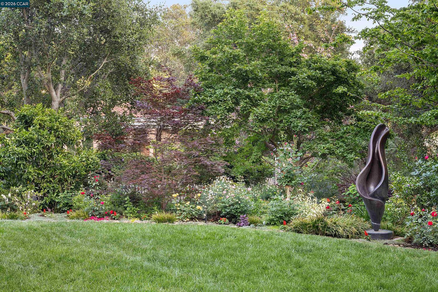 This lush, well-manicured garden features a vibrant mix of mature trees, flowering shrubs, and a neatly trimmed lawn. A striking, dark-toned abstract sculpture stands prominently on the right side, serving as a focal point amidst the colorful floral beds. The scene offers a serene and private atmosphere, perfect for outdoor relaxation or garden enthusiasts.